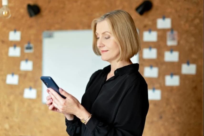 Kobieta ubrana w czarną bluzkę trzyma w dłoniach telefon komórkowy.